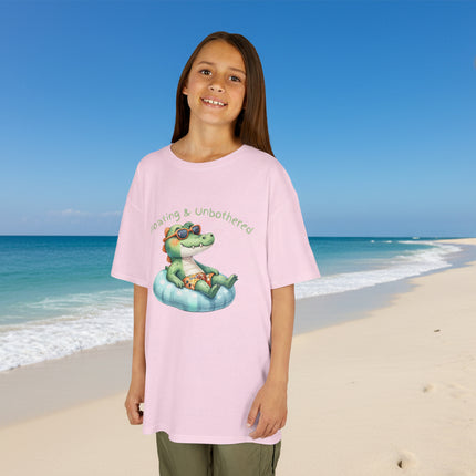 Young girl wearing a pink t-shirt with a graphic of a crocodile on an inflatable ring and text 'Unbothered & Unbothered'.